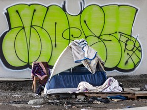There is bright green grafitti behind an unhoused person's tent and chair on urban gravel.