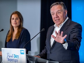 Francois Legault, in a blue suit, holds his hand up as he gestures at a podium. Pascale Dery, also in a dark suit, is in the background.