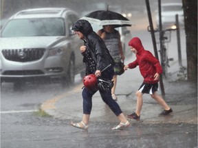 Two people are walking across the street while getting drenched from the rain. A woman wears a black raincoat and the son wears a red one.