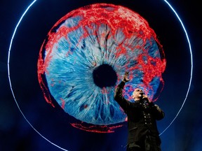 Peter Gabriel sings with one arm raised. In the background, a giant blue and red eye on a screen