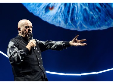 Peter Gabriel with his arm extended as he sings in front of a blue and neon visual.
