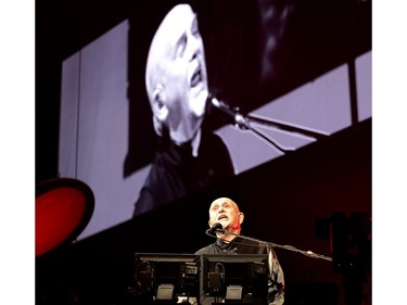 Peter Gabriel performs sitting down, with a large screen behind him showing him singing.