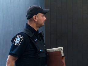 A police officer in a cap and uniform holds a folder as he walks in a courtroom.