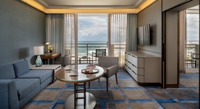 The sitting area of a suite with sofa and table. It has blue and grey accents and overlooks the Mediterranean Sea.