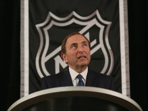 Commissioner Gary Bettman, in a dark suit with a white shirt and a black tie, stands in front of the NHL logo.
