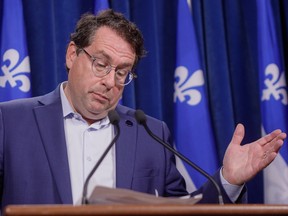 Bernard Drainville gestures in front of a podium with Quebec flags behind him