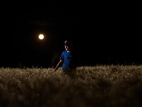 A boy wearing a ballcap stands smiling in a wheat field. The full moon is behind him.