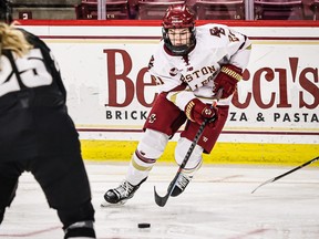 Abby Newhook, recently named co-captain of Boston College's women's hockey team, is seen in a white jersey and red pants carrying the puck during a game.