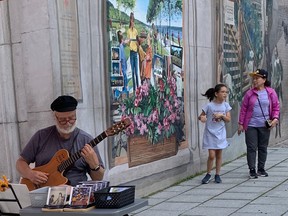 A woman and girl walk past a Quebec City mural toward a man selling CDs on the street. The man is playing a guitar.