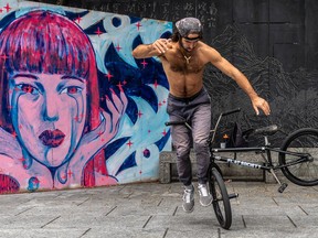 A shirtless man does a trick with a bicycle in front of a mural of a woman with pink hair