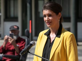 Genevieve Guilbault in a bright yellow blazer speaks during a press conference. Behind her is a man in a wheelchair taking a photo with his phone.