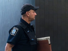 A police officer is seen from the side profile holding a file beneath his arm. His emblem for the Montreal police is visible on his right arm as he stares towards the right side of the frame, with a baseball cap on.