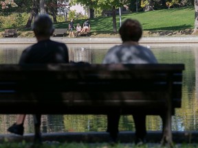 Two people are seen from behind on a park bench. They are looking across a pond at three people in shorts and t-shirts chatting on another bench.