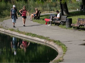 Two people stroll along a path by a pond. There are people sitting on benches facing the water.