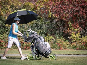 A woman pushes a golf trolley while holding an umbrella.