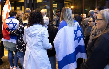 People wait to enter a building for a vigil marking the deaths of over 800 Israelis in a Hamas attack. Several people are draped in Israeli flags.