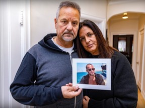 A woman leans her head on the shoulder of a man, who is holding up a picture of their adult son wearing sunglasses by the water
