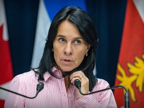 Montreal Mayor Valérie Plante, wearing a pink and white striped shirt, speaks into microphones in front of the Quebec, Montreal and Canadian flags.