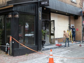 Two workers place plywood over a broken window on the facade of a restaurant. Police tape blocks off the scene. A sign on the side of the building reads "Meze avec moi."