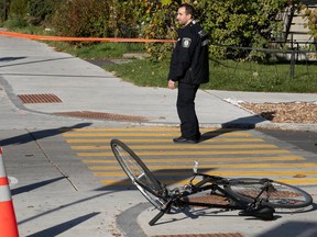A bicycle lies on a sidewalk at a street corner surrounded by orange police tape with an officer standing nearby