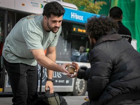 A young bearded Montreal police recruit hands food to an unhoused man as a city bus drives by.