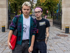 Two men are standing in front of school gates. One is wearing a cardigan over a t-shirt and has blonde hair. The other is wearing a t-shirt, glasses and a bandana. Both are staring directly at the camera, standing straight in front of it.