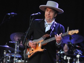 Bob Dylan, wearing a suit and brimmed hat, plays guitar on stage.