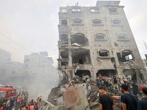 A bombed out building seen through the haze of smoke. there is a fire truck at bottom left and people gathered to look at and go through the wreckage.