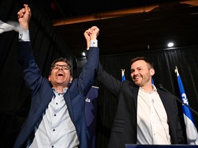 Two men hold their hands high in victory in front of the Quebec flag.
