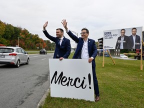 Paul St-Pierre Plamondon and Pascal Paradis hold a large sign that says "Merci!" on the side of a road. Behind them is a large election sign featuring them.