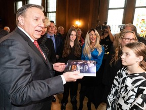 Quebec Premier François Legault talks to a gathering while holding a copy of a book about hockey player Mike Bossy. A child watches him speak to someone off-camera.