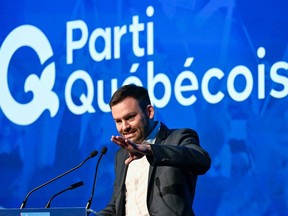 Paul St-Pierre Plamondon gestures speaking at a microphone with a giant Parti Québécois logo on a screen behind him
