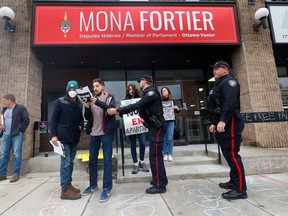 Protesters and police stand outside the office of Mona Fortier. Messages are written in chalk on the sidewalk.