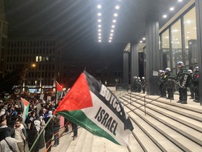 Police in riot gear stand guard at the top of steps leading to a large building while protesters carrying Palestinian flags gather below