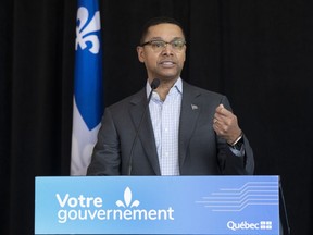 A man speaks at a podium with a sign reading "Votre gouvernement" and a Quebec flag in the background.