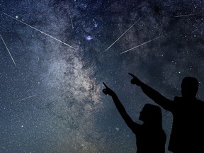 Two people are silhouetted against a night sky. They are pointing toward a meteor shower.