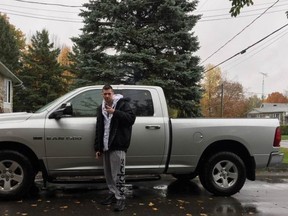 A man is standing in front of a grey pickup truck, ironically throwing up a peace sign.