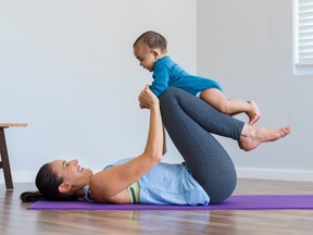 A woman on her back on a yoga mat rests her baby on her shins to do leg lifts.