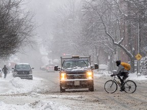 A truck drives on a snowy road with its headlights on. There is a pedestrian on the sidewalk and a cyclist crossing the road in front of the truck.
