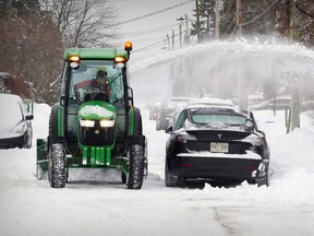 A green snowplow driving between cars blows snow from a road toward a sidewalk.