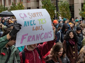 A large group of university students march down a street during a protest. Two students in the foreground are holding a sign reading "Ça suffit avec tes lois, François."