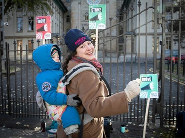 Anna Lewton-Brain, carrying a child on her back, smiles as she looks over her shoulder at the camera, outside a locked fence gate with strike posters attached to it. On her head is a tuque that reads 'Profs en négo'