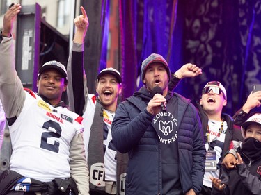 Jason Maas speaks into a microphone while Alouettes players cheer behind him on stage