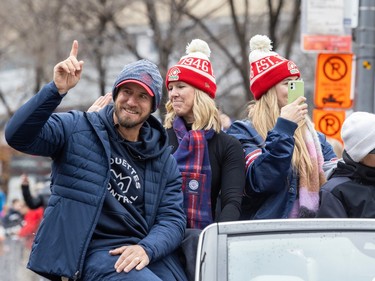 Jason Maas points a finger in the air and smiles as he rides in the back of an open-top car with two women during a parade