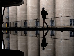 A man jogging in an industrial area with large silos is reflected in the water of a canal.