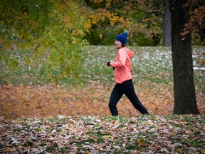 A woman in a pink windbreaker and blue hat jobs on a path with fallen leaves and a little snow.
