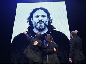 Three people wave to a large image of Karl Tremblay on a screen on stage