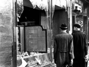 In a black and white photo, two men walk past a broken shop window.