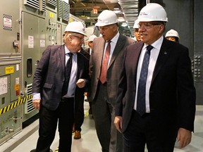 Jean Charest, Pierre Fitzgibbon and François Legault, wearing hard hats, walk by large machines on a plant floor