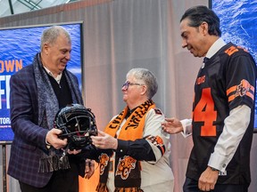 Randy Ambrosie hands a helmet to Marianne Alto, who is wearing a Lions scarf, next to Amar Doman in a Lions jersey